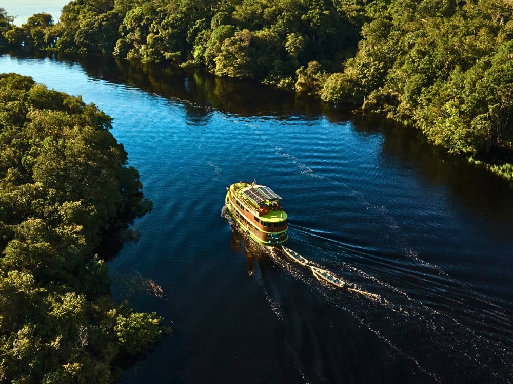 Veduta aerea di una barca da crociera che naviga su un fiume scuro dell'Amazzonia, circondato dalla fitta foresta verde, in Brasile. Veduta aerea di una barca da crociera che naviga su un fiume scuro dell'Amazzonia, circondato dalla fitta foresta verde, in Brasile.