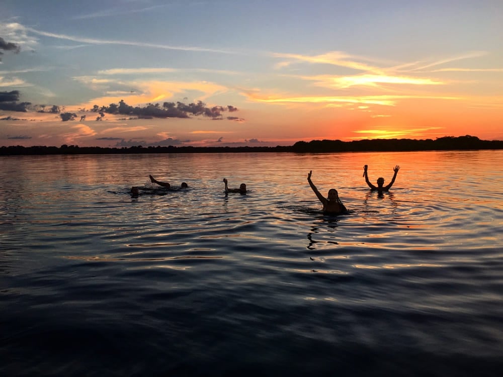 Un gruppo di viaggiatori nuota in un fiume di acque nere dell'Amazzonia durante un tramonto arancione acceso. Un gruppo di viaggiatori nuota in un fiume di acque nere dell'Amazzonia durante un tramonto arancione acceso.