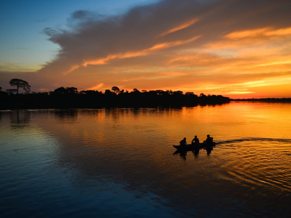 Viaggio nella foresta amazzonica: la sagoma di tre persone su una piccola canoa che naviga sul Rio delle Amazzoni contro un cielo arancione al tramonto. Viaggio nella foresta amazzonica: la sagoma di tre persone su una piccola canoa che naviga sul Rio delle Amazzoni contro un cielo arancione al tramonto.