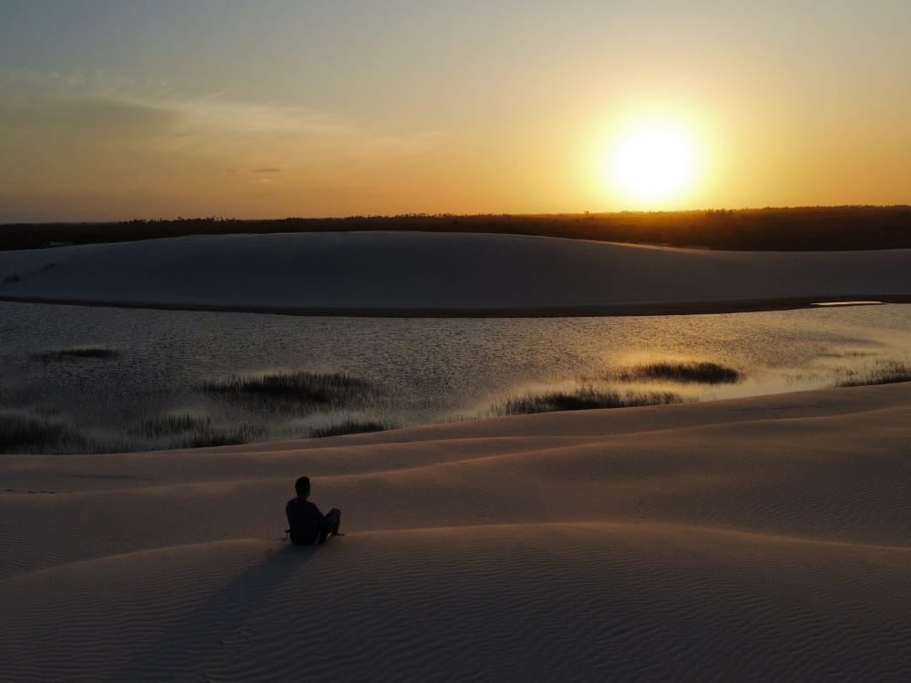 Silueta de una persona sentada en una duna de arena mientras contempla un atardecer dorado sobre las lagunas de los Lençóis Maranhenses. Silueta de una persona sentada en una duna de arena mientras contempla un atardecer dorado sobre las lagunas de los Lençóis Maranhenses.