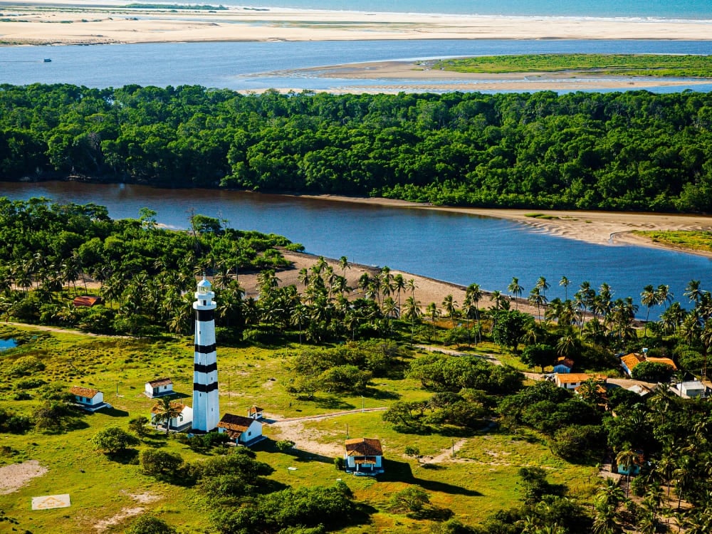 Veduta aerea di un faro alto a strisce bianche e nere e di una vegetazione verde e rigogliosa vicino alle città base dei tour nei Lençóis Maranhenses. Veduta aerea di un faro alto a strisce bianche e nere e di una vegetazione verde e rigogliosa vicino alle città base dei tour nei Lençóis Maranhenses.