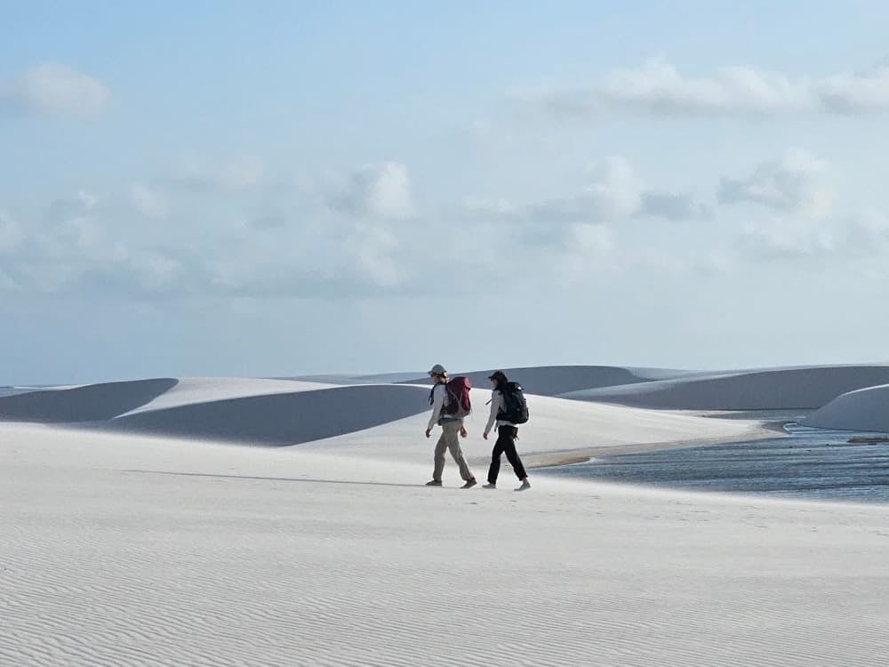 Due escursionisti con lo zaino attraversano le grandi dune di sabbia bianca accanto a una laguna d'acqua dolce, nel Parco Nazionale dei Lençóis Maranhenses. Due escursionisti con lo zaino attraversano le grandi dune di sabbia bianca accanto a una laguna d'acqua dolce, nel Parco Nazionale dei Lençóis Maranhenses.