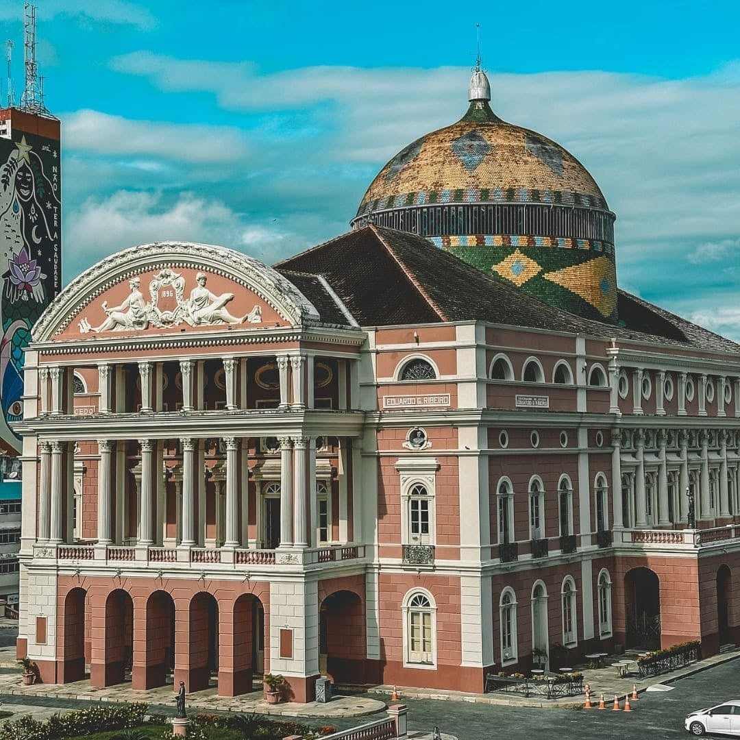 La facciata neoclassica rosa e la cupola di piastrelle colorate del Teatro Amazonas di Manaus, sotto un cielo azzurro. La facciata neoclassica rosa e la cupola di piastrelle colorate del Teatro Amazonas di Manaus, sotto un cielo azzurro.