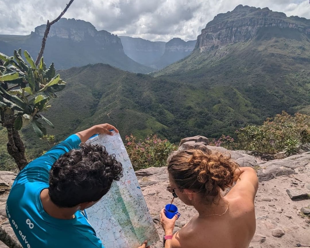 Una guida locale di PlanetaEXO con la camicia blu mostra la mappa dei sentieri a un'escursionista, con una grande valle verde e le montagne sullo sfondo. Una guida locale di PlanetaEXO con la camicia blu mostra la mappa dei sentieri a un'escursionista, con una grande valle verde e le montagne sullo sfondo.