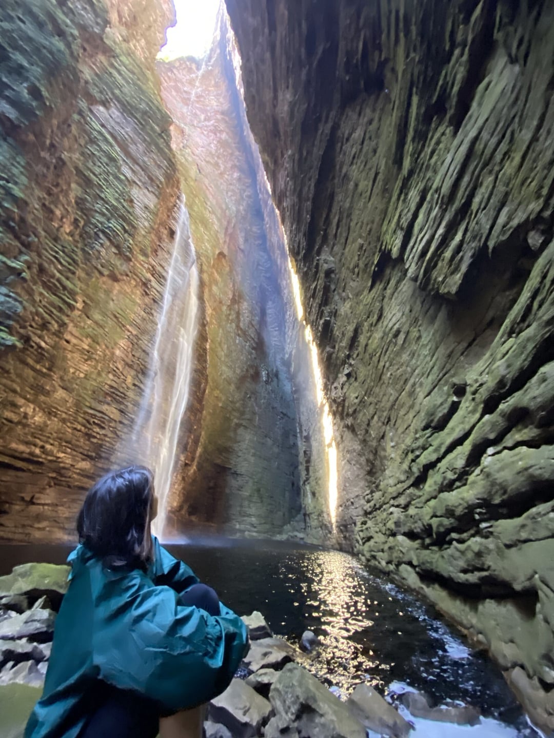 A hiker in a green jacket admires a towering waterfall inside a steep canyon during the rainy season in Chapada Diamantina. A hiker in a green jacket admires a towering waterfall inside a steep canyon during the rainy season in Chapada Diamantina.