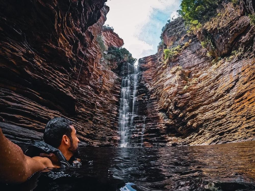 Un uomo nuota in una piscina naturale scura e guarda verso l'alto una cascata altissima che scende lungo un canyon di roccia stratificata. Un uomo nuota in una piscina naturale scura e guarda verso l'alto una cascata altissima che scende lungo un canyon di roccia stratificata.