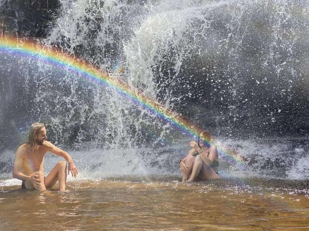 Due viaggiatori seduti nell'acqua bassa ai piedi di una cascata, con il riflesso di un arcobaleno. Due viaggiatori seduti nell'acqua bassa ai piedi di una cascata, con il riflesso di un arcobaleno.