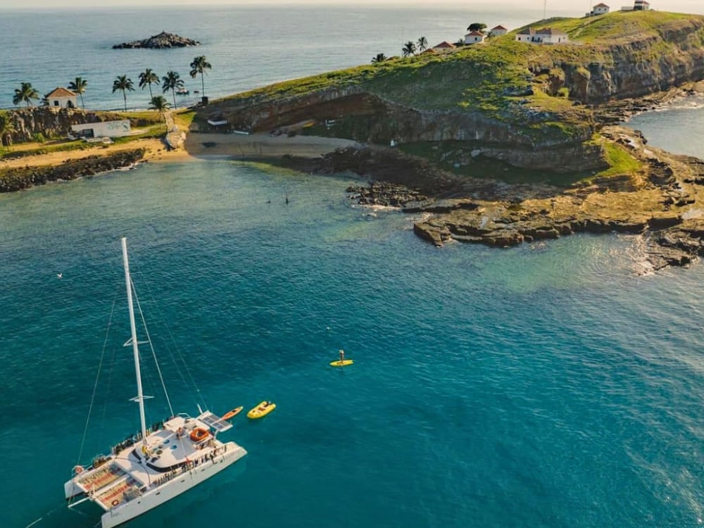 Luftaufnahme eines weißen Katamarans, der in kristallklarem Wasser neben einer grünen Insel im Abrolhos-Archipel ankert. Luftaufnahme eines weißen Katamarans, der in kristallklarem Wasser neben einer grünen Insel im Abrolhos-Archipel ankert.