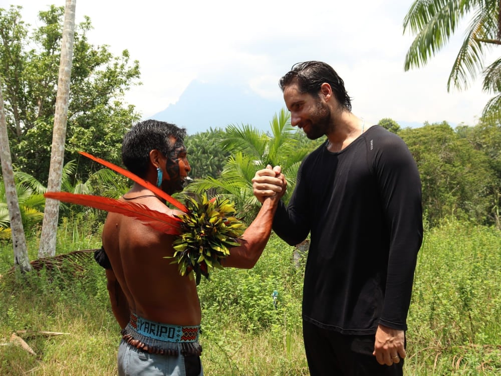 Una guida indigena yanomami con le tradizionali piume rosse stringe la mano a un escursionista nella foresta. Una guida indigena yanomami con le tradizionali piume rosse stringe la mano a un escursionista nella foresta.