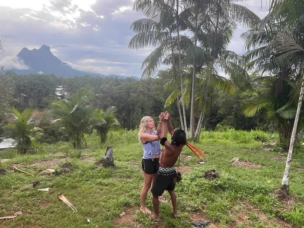 Katharina che interagisce in modo rilassato con una guida indigena Yanomami in una zona verde, con il Pico da Neblina sullo sfondo. Katharina che interagisce in modo rilassato con una guida indigena Yanomami in una zona verde, con il Pico da Neblina sullo sfondo.