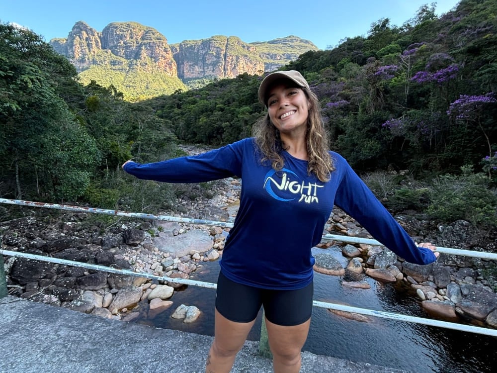 Mariana sorride con le braccia aperte su un ponte sopra un fiume di rocce, con le montagne aspre del Vale do Pati sullo sfondo. Mariana sorride con le braccia aperte su un ponte sopra un fiume di rocce, con le montagne aspre del Vale do Pati sullo sfondo.