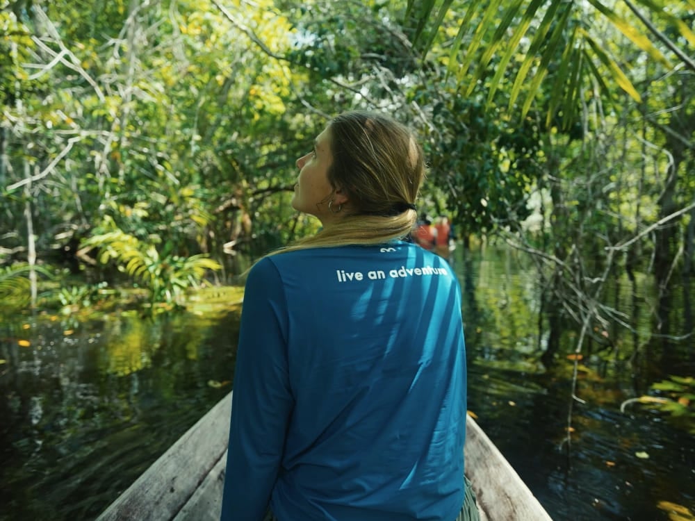 Donna di spalle su una barca di legno che naviga un fiume circondato da foresta fitta, con una maglietta blu con la scritta live an adventure. Donna di spalle su una barca di legno che naviga un fiume circondato da foresta fitta, con una maglietta blu con la scritta live an adventure.