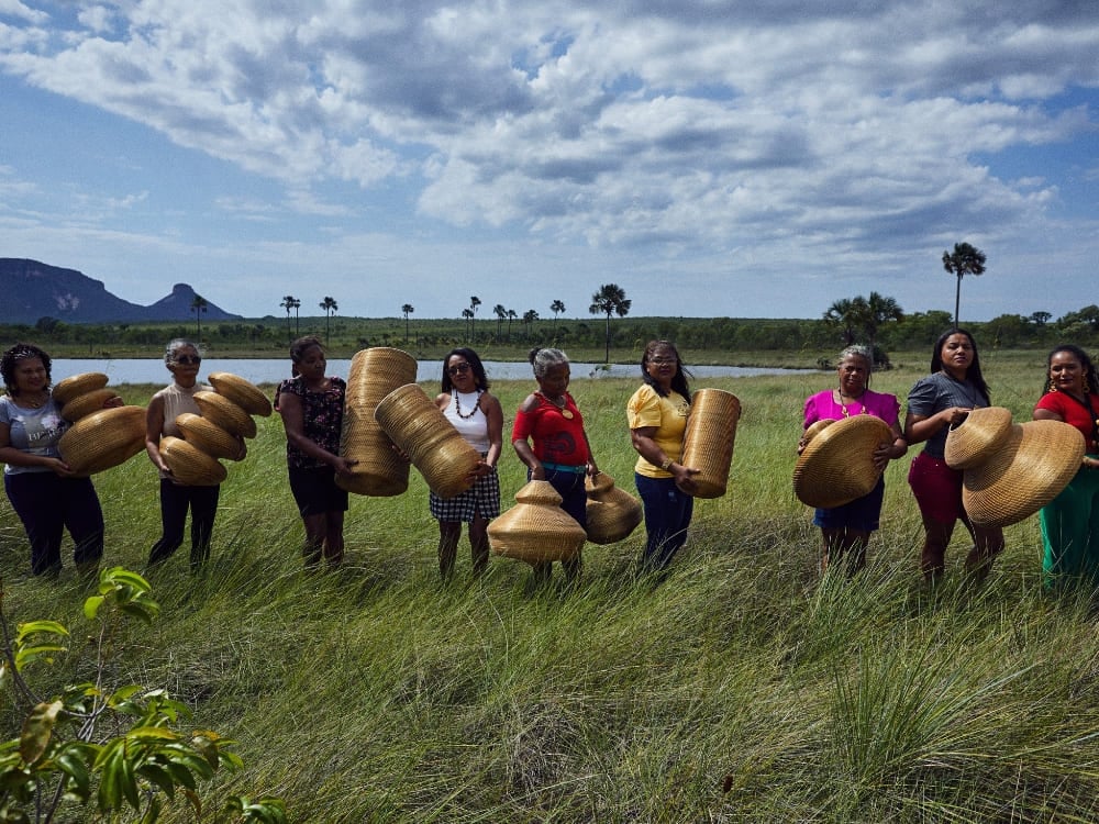 Um grupo de mulheres de pé em um campo verde exuberante sob um céu nublado, segurando artesanatos tecidos feitos de Capim Dourado na região do Jalapão. Um grupo de mulheres de pé em um campo verde exuberante sob um céu nublado, segurando artesanatos tecidos feitos de Capim Dourado na região do Jalapão.