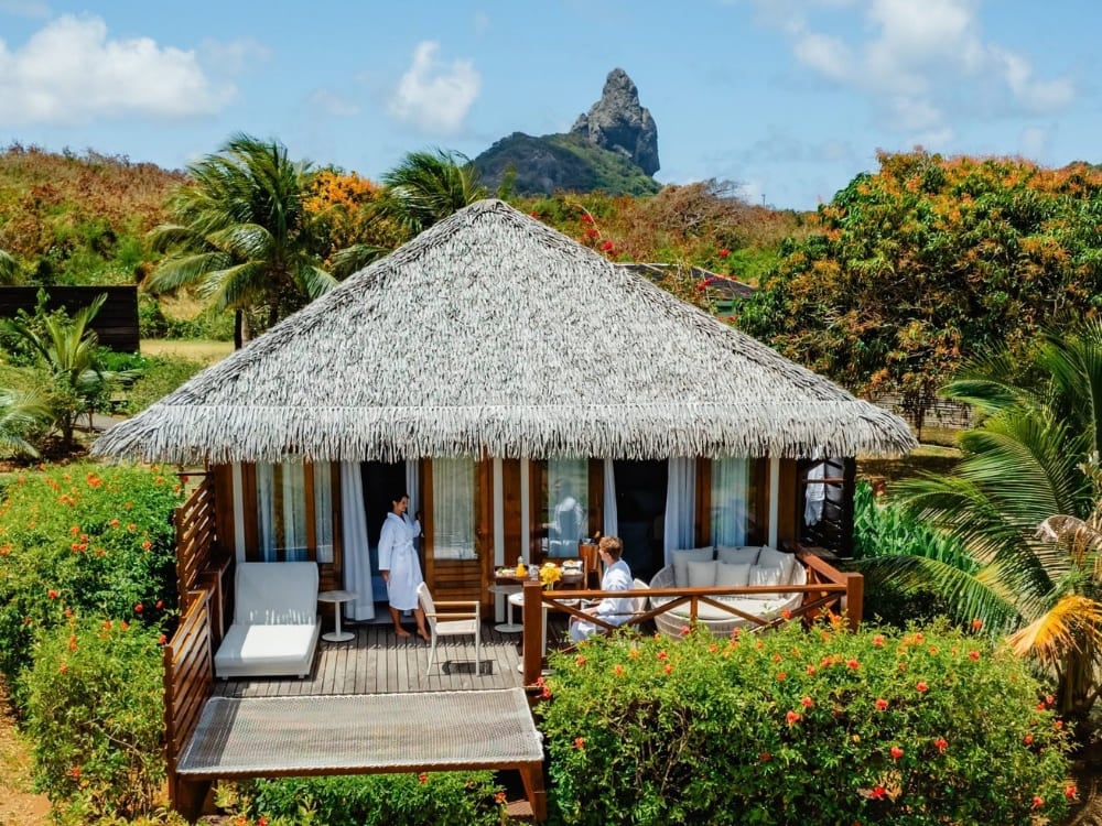 Coppia in accappatoio bianco che si rilassa sul deck privato di un bungalow rustico con tetto di paglia e vista sul Morro do Pico, in uno dei migliori hotel di Fernando de Noronha. Coppia in accappatoio bianco che si rilassa sul deck privato di un bungalow rustico con tetto di paglia e vista sul Morro do Pico, in uno dei migliori hotel di Fernando de Noronha.