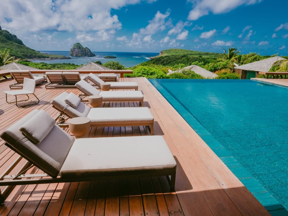 Deck in legno con comode sedie a sdraio accanto a una piscina a sfioro affacciata sull’oceano e sul Morro do Pico, in un hotel di Fernando de Noronha Deck in legno con comode sedie a sdraio accanto a una piscina a sfioro affacciata sull’oceano e sul Morro do Pico, in un hotel di Fernando de Noronha