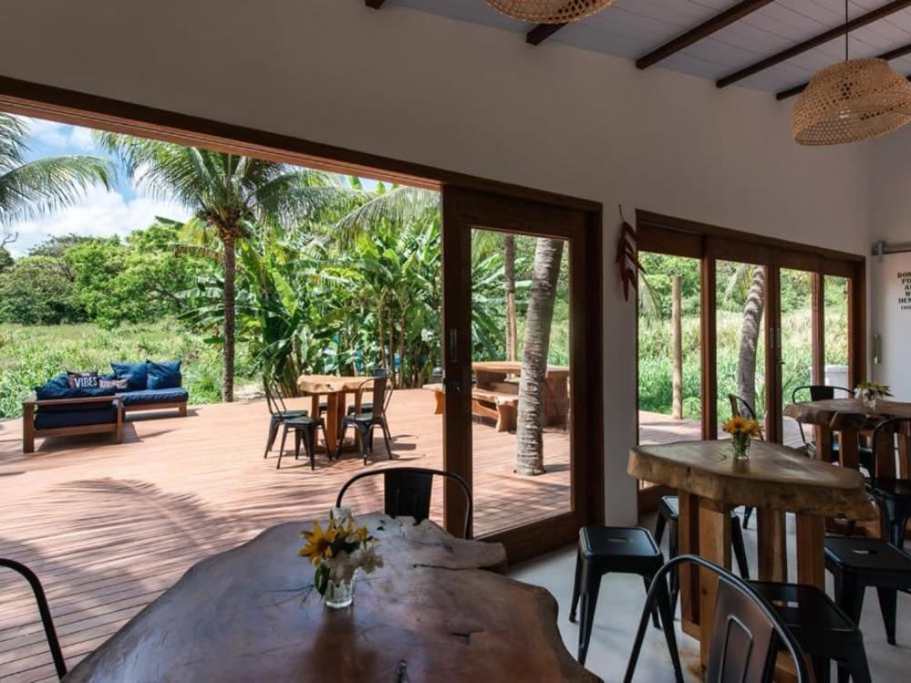 Area comune all’aperto di un ostello, con un tavolo rustico in legno e ampie porte affacciate sulla vegetazione rigogliosa di Fernando de Noronha. Area comune all’aperto di un ostello, con un tavolo rustico in legno e ampie porte affacciate sulla vegetazione rigogliosa di Fernando de Noronha.