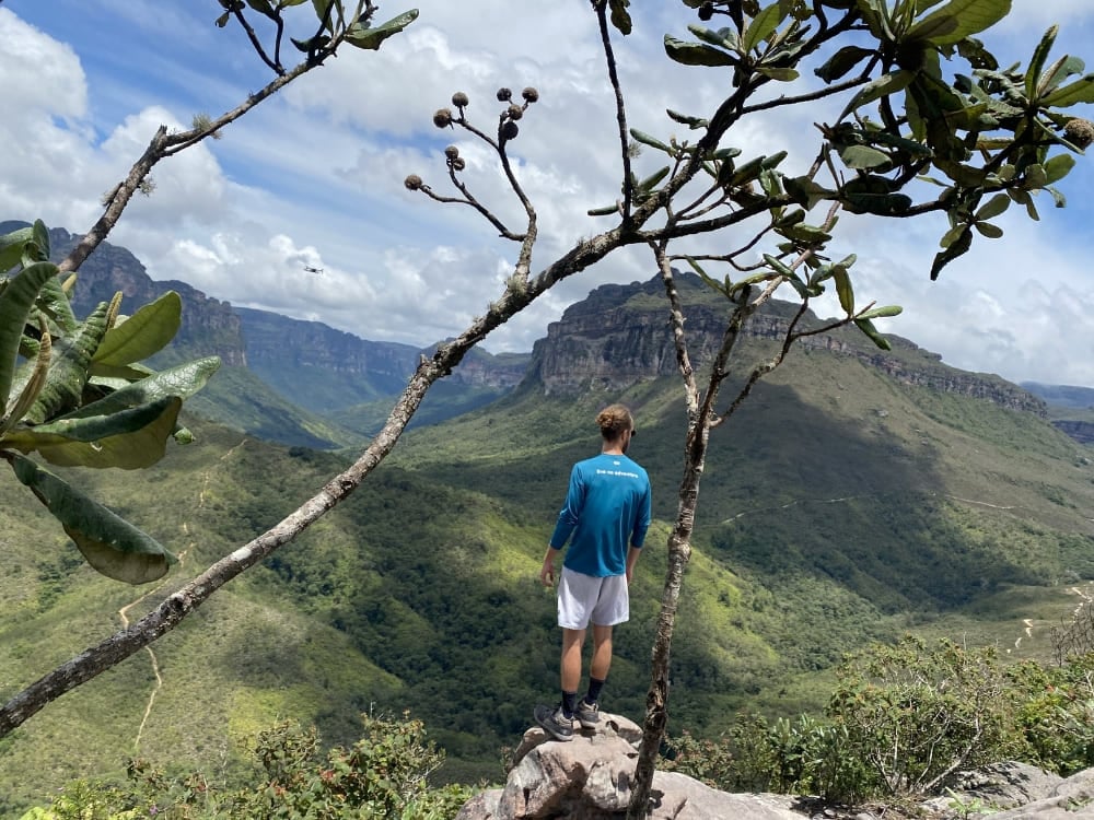 Een wandelaar staat op een rotsrand omringd door boomtakken en kijkt uit over de groene vallei naar de platte top van Morro do Castelo. Een wandelaar staat op een rotsrand omringd door boomtakken en kijkt uit over de groene vallei naar de platte top van Morro do Castelo.