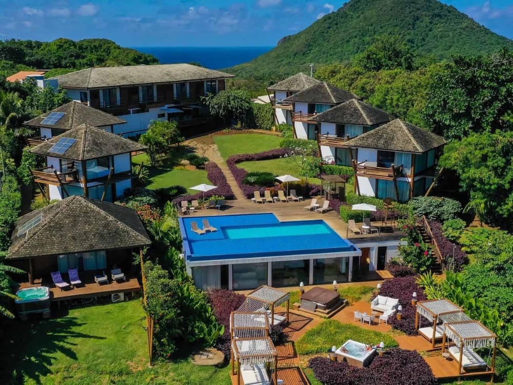 Veduta aerea di Pousada Morena con i bungalow dal tetto scuro, la piscina blu centrale e le vasche idromassaggio circondate dalla foresta rigogliosa di Fernando de Noronha. Veduta aerea di Pousada Morena con i bungalow dal tetto scuro, la piscina blu centrale e le vasche idromassaggio circondate dalla foresta rigogliosa di Fernando de Noronha.