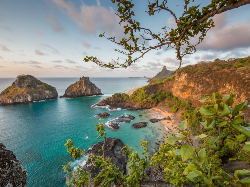 Scenic coastal view of Fernando de Noronha featuring turquoise waters, a sandy cove, and the Morro Dois Irmãos rock formations framed by green vegetation. Scenic coastal view of Fernando de Noronha featuring turquoise waters, a sandy cove, and the Morro Dois Irmãos rock formations framed by green vegetation.