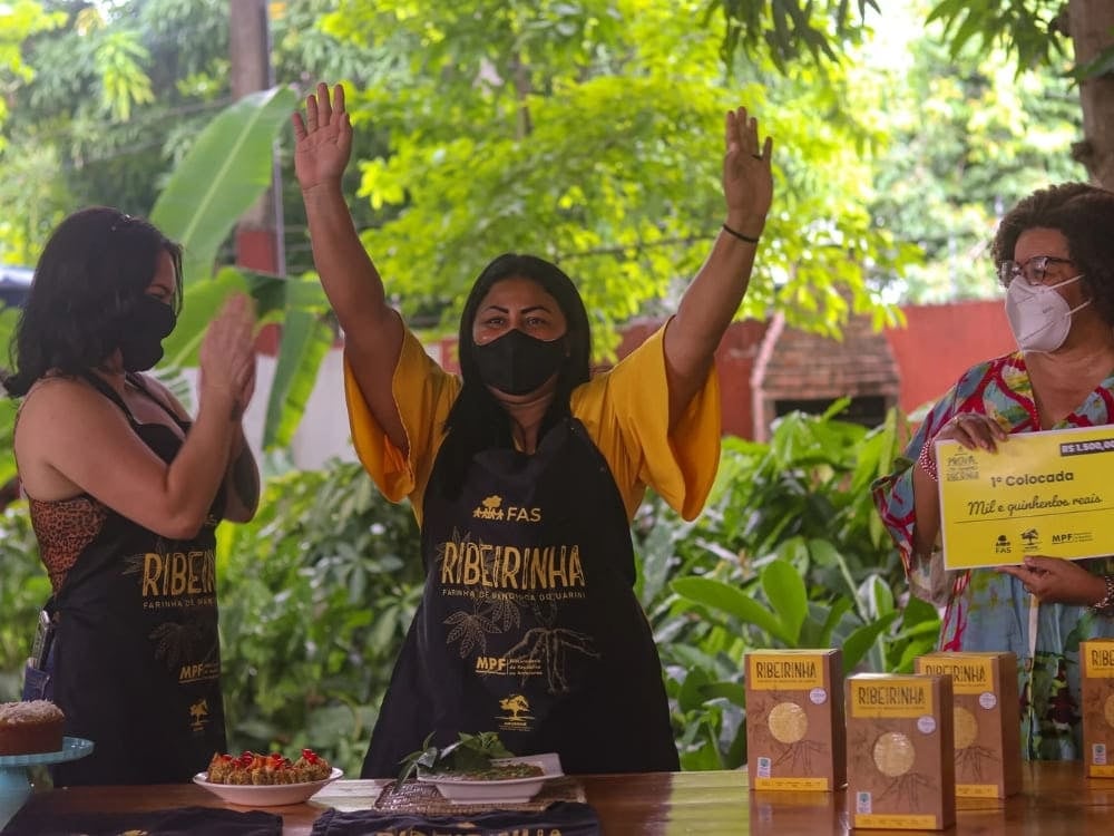 Uma mulher usando um avental preto com a marca FAS Ribeirinha levanta as mãos de alegria, ao lado de outras pessoas celebrando o programa da Fundação Amazônia Sustentável. Uma mulher usando um avental preto com a marca FAS Ribeirinha levanta as mãos de alegria, ao lado de outras pessoas celebrando o programa da Fundação Amazônia Sustentável.