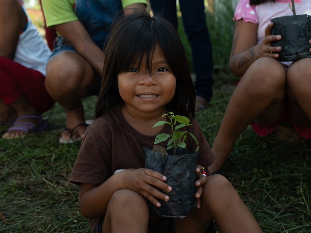 Un niño sonriente de una comunidad ribereña amazónica sentado en la hierba y sosteniendo una pequeña planta en una maceta. Un niño sonriente de una comunidad ribereña amazónica sentado en la hierba y sosteniendo una pequeña planta en una maceta.