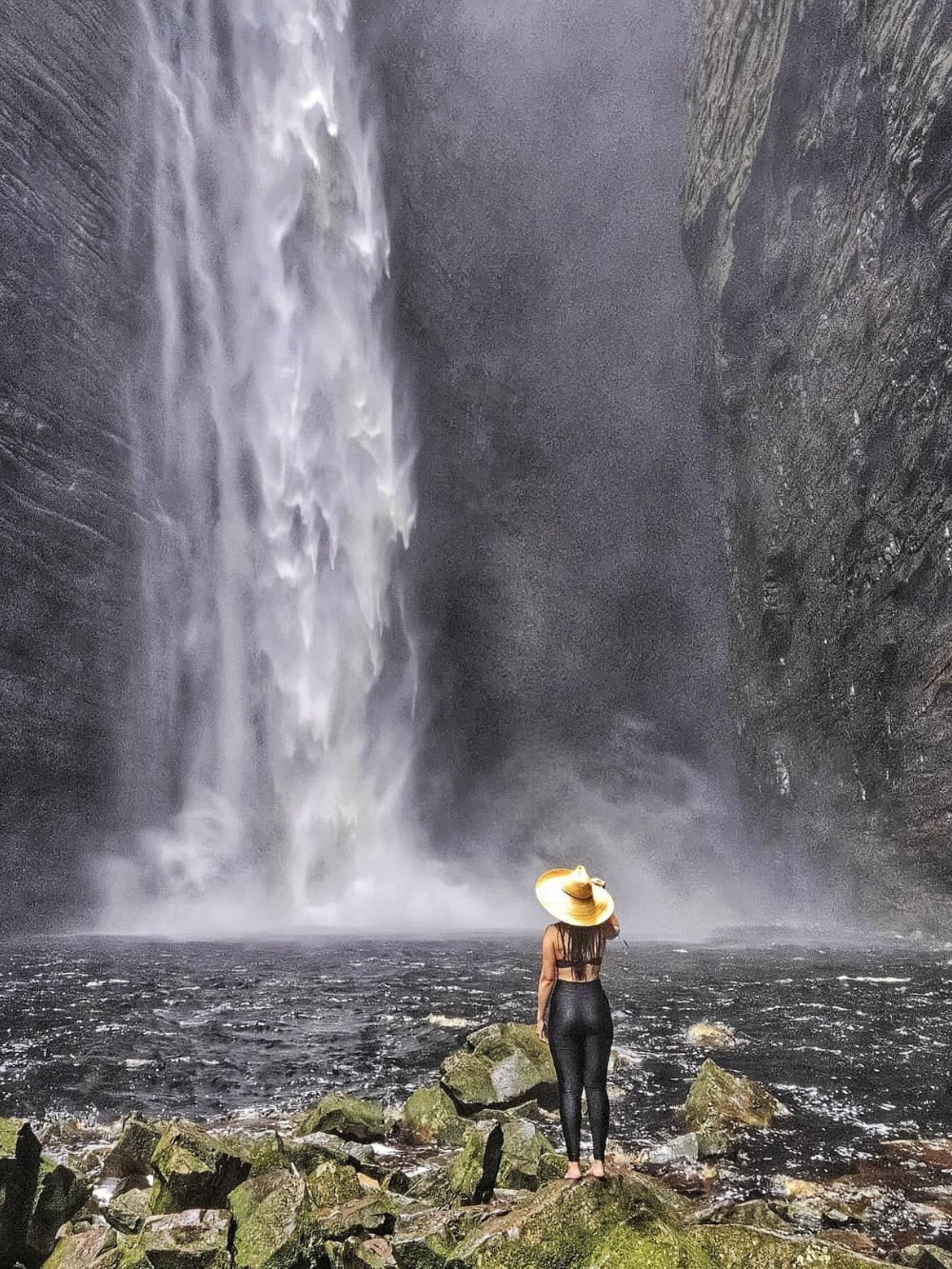 Un escursionista con il cappello sul bordo di una pozza scura guarda il salto verticale di 100 metri della cascata da Fumacinha, dentro un canyon stretto. Un escursionista con il cappello sul bordo di una pozza scura guarda il salto verticale di 100 metri della cascata da Fumacinha, dentro un canyon stretto.