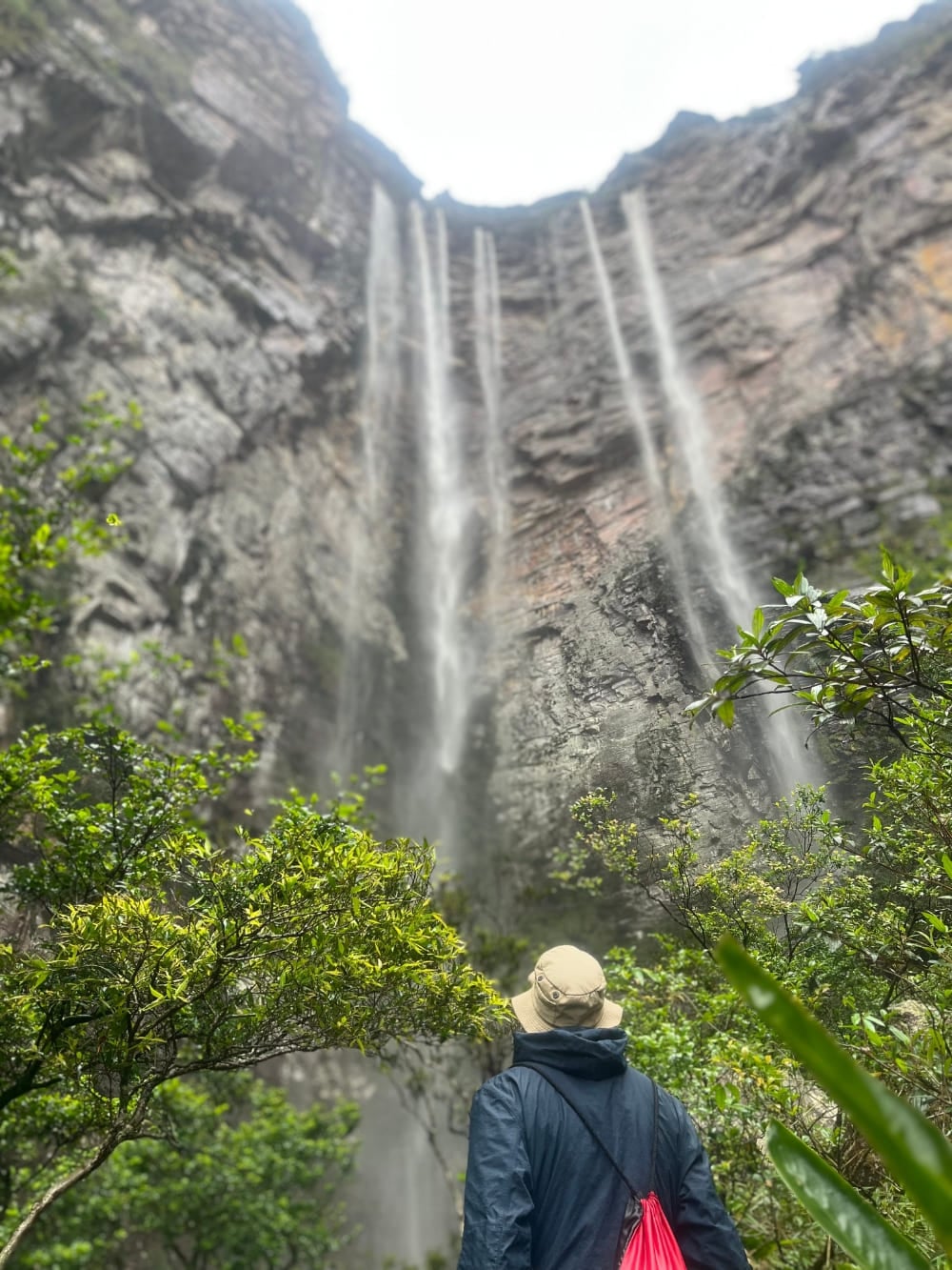 Un escursionista con cappello e giacca osserva dalla base rocciosa i getti d'acqua di 280 metri del Cachoeirão. Un escursionista con cappello e giacca osserva dalla base rocciosa i getti d'acqua di 280 metri del Cachoeirão.
