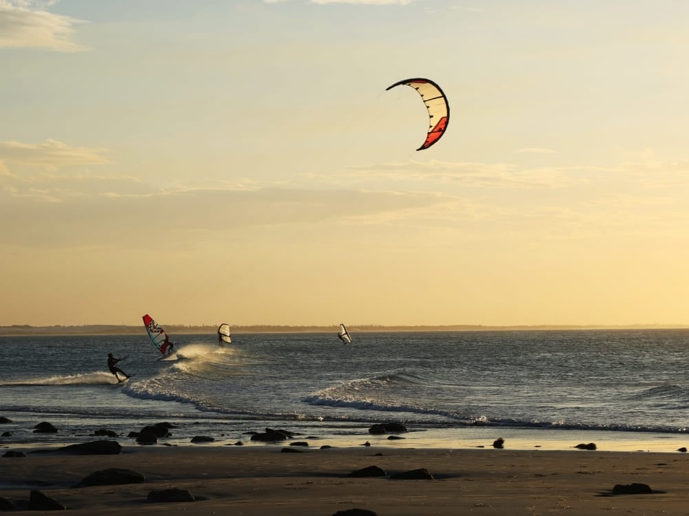 Silhouettes of people kitesurfing and windsurfing on ocean waves near a sandy beach during a golden sunset. Silhouettes of people kitesurfing and windsurfing on ocean waves near a sandy beach during a golden sunset.