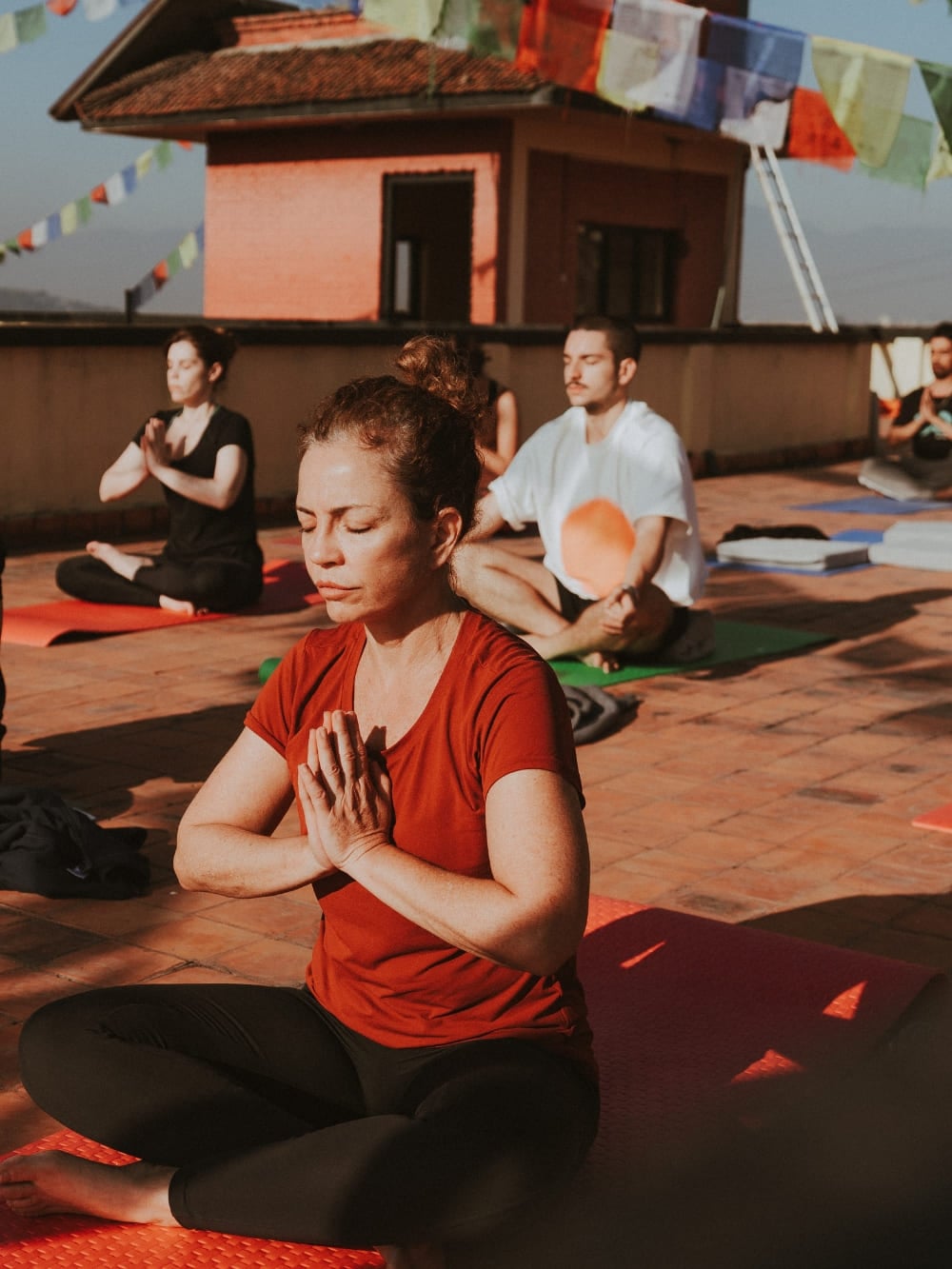 Mila Ricetti und Lorenzo praktizieren Yoga und meditieren mit einer Gruppe auf einem sonnendurchfluteten Dach, das mit Gebetsfahnen geschmückt ist, während eines Retreats in Nepal. Mila Ricetti und Lorenzo praktizieren Yoga und meditieren mit einer Gruppe auf einem sonnendurchfluteten Dach, das mit Gebetsfahnen geschmückt ist, während eines Retreats in Nepal.