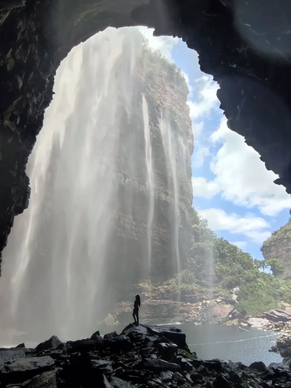 La sagoma di una persona in piedi sulle rocce vicino all'ingresso di una grotta scura guarda le pareti irregolari di 100 metri della cascata do Herculano. La sagoma di una persona in piedi sulle rocce vicino all'ingresso di una grotta scura guarda le pareti irregolari di 100 metri della cascata do Herculano.