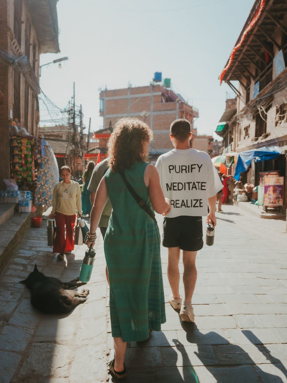 Mila und Lorenzo gehen Seite an Seite eine sonnige Straße in Nepal hinunter, wobei Lorenzo ein T-Shirt mit der Aufschrift „Purify, Meditate, Realize“ trägt. Mila und Lorenzo gehen Seite an Seite eine sonnige Straße in Nepal hinunter, wobei Lorenzo ein T-Shirt mit der Aufschrift „Purify, Meditate, Realize“ trägt.