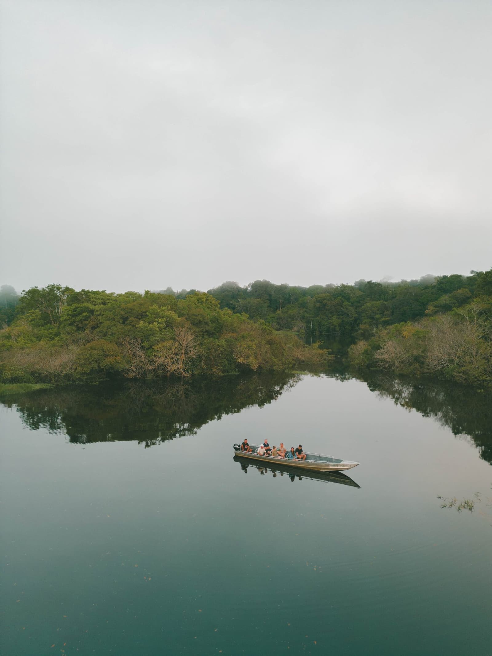 Sechs Passagiere, ein Fluss, und der Wald hält ringsum den Atem an. Ein kleines Motorboot mit einer Reisegruppe gleitet auf einem spiegelglatten, nebelverhangenen Fluss bei einem Ausflug einer Amazon Boutique Lodge, umgeben von dichtem Tropenwald, der sich im stillen Wasser spiegelt