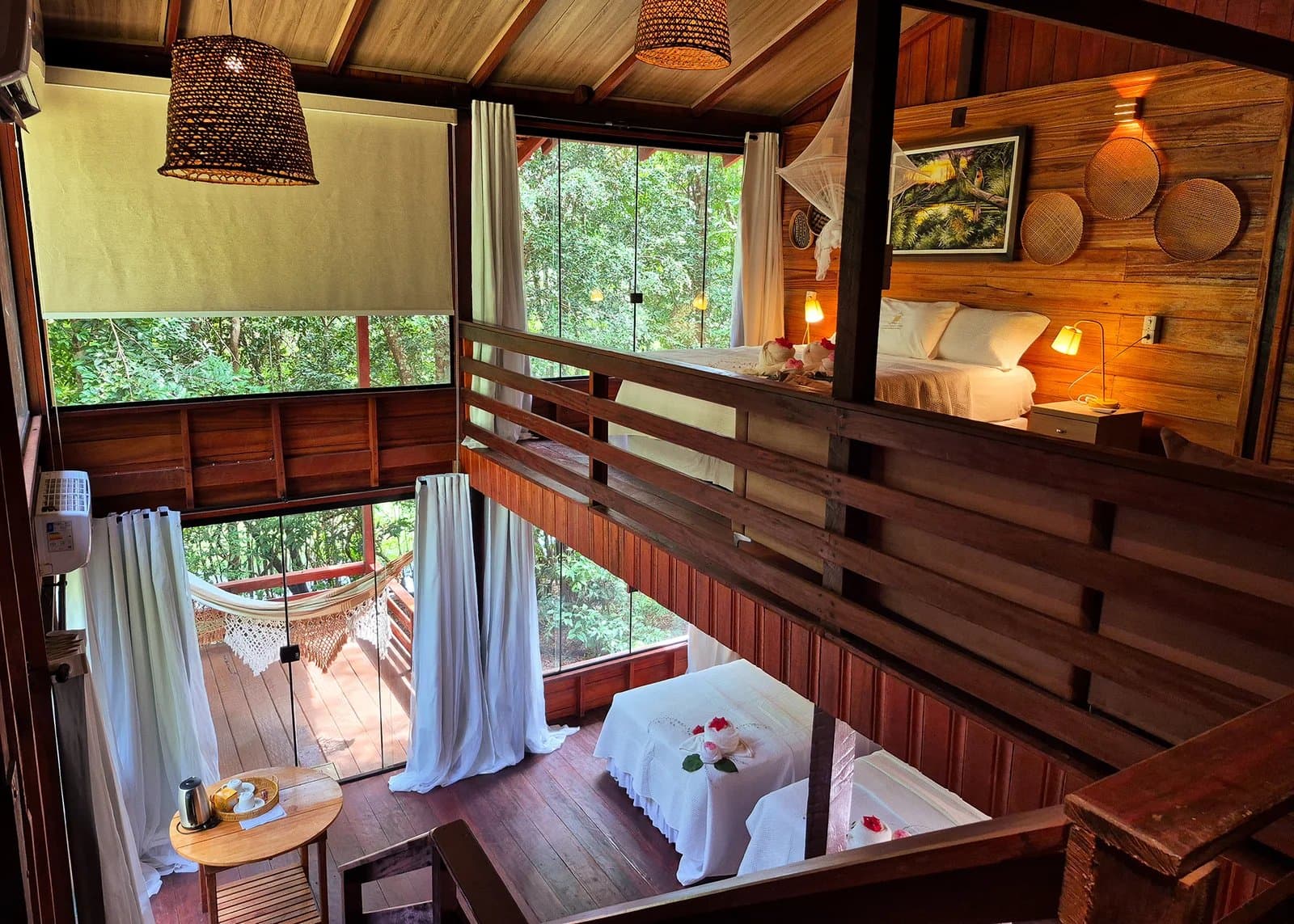 Rosenblätter, Hängematte, Regenwald. Mehr braucht es nicht. Blick von der Galerie eines Bungalows in einer Amazon-Boutique-Lodge mit warmem Holzinterieur, weißer Bettwäsche, geflochtenen Pendelleuchten und raumhohen Fenstern zum dichten Regenwald, darunter ein Wohnbereich mit Massagetisch, zarten weißen Vorhängen und einer Hängematte auf der Veranda