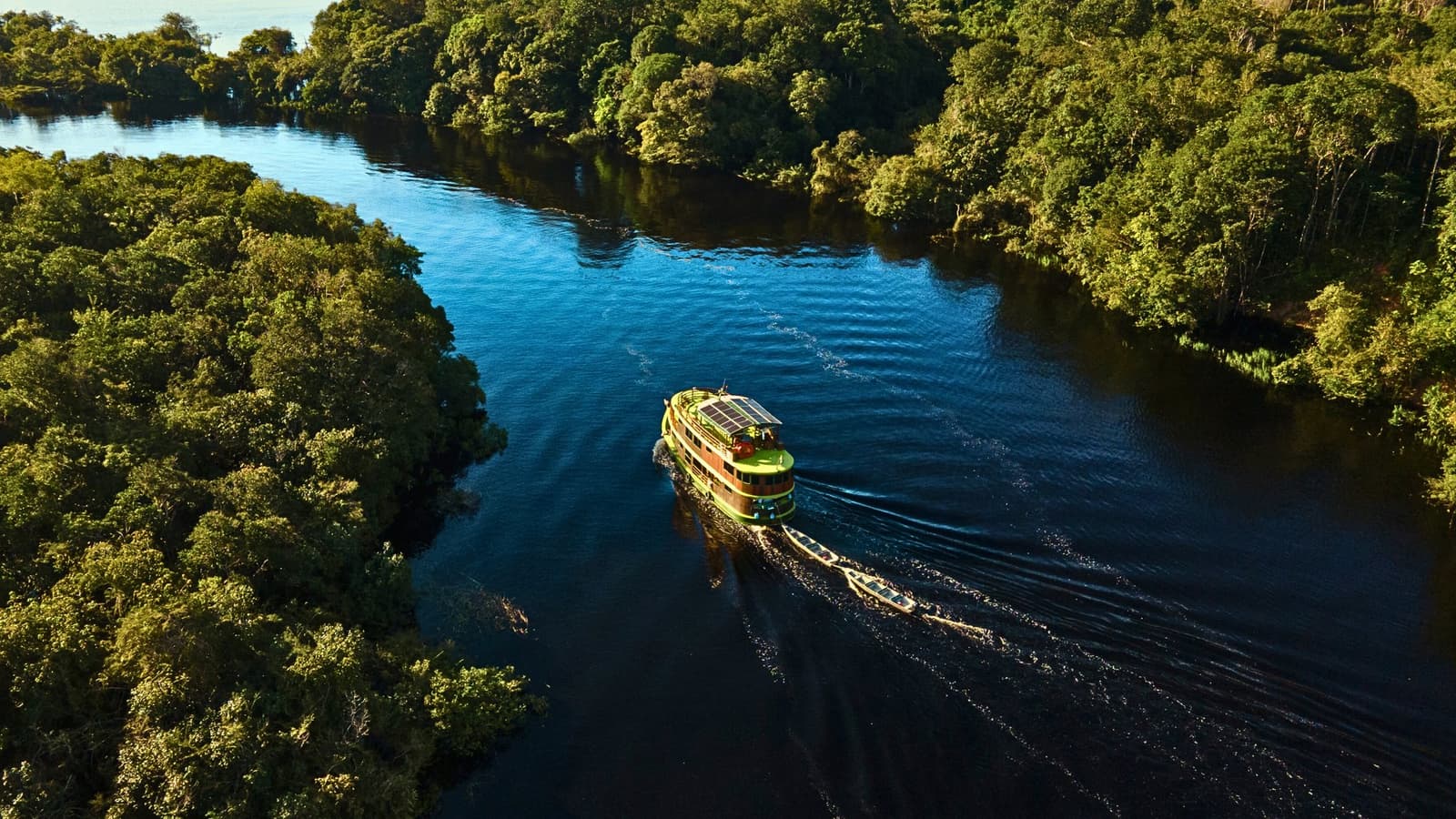 Regenwald rechts, Regenwald links, und das Schiff mittendrin. Luftaufnahme eines Amazon-Flusskreuzfahrtboots mit Solarpanelen, das mit einem kleinen Beiboot einen dunklen Schwarzwasserfluss durch dichten Regenwald navigiert