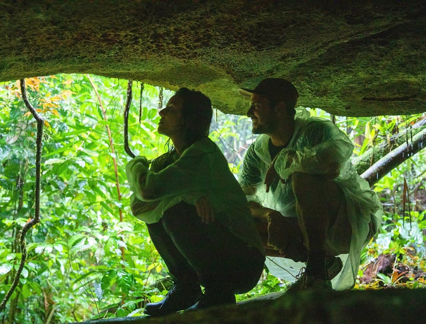 Regen mitten im Hike? Der Regenwald kennt eben seinen eigenen Rhythmus. Zwei Reisende hocken unter einem moosbedeckten Felsvorsprung im dichten Regenwald, in Regenponchons gekleidet und von üppigem grünem Blattwerk umgeben