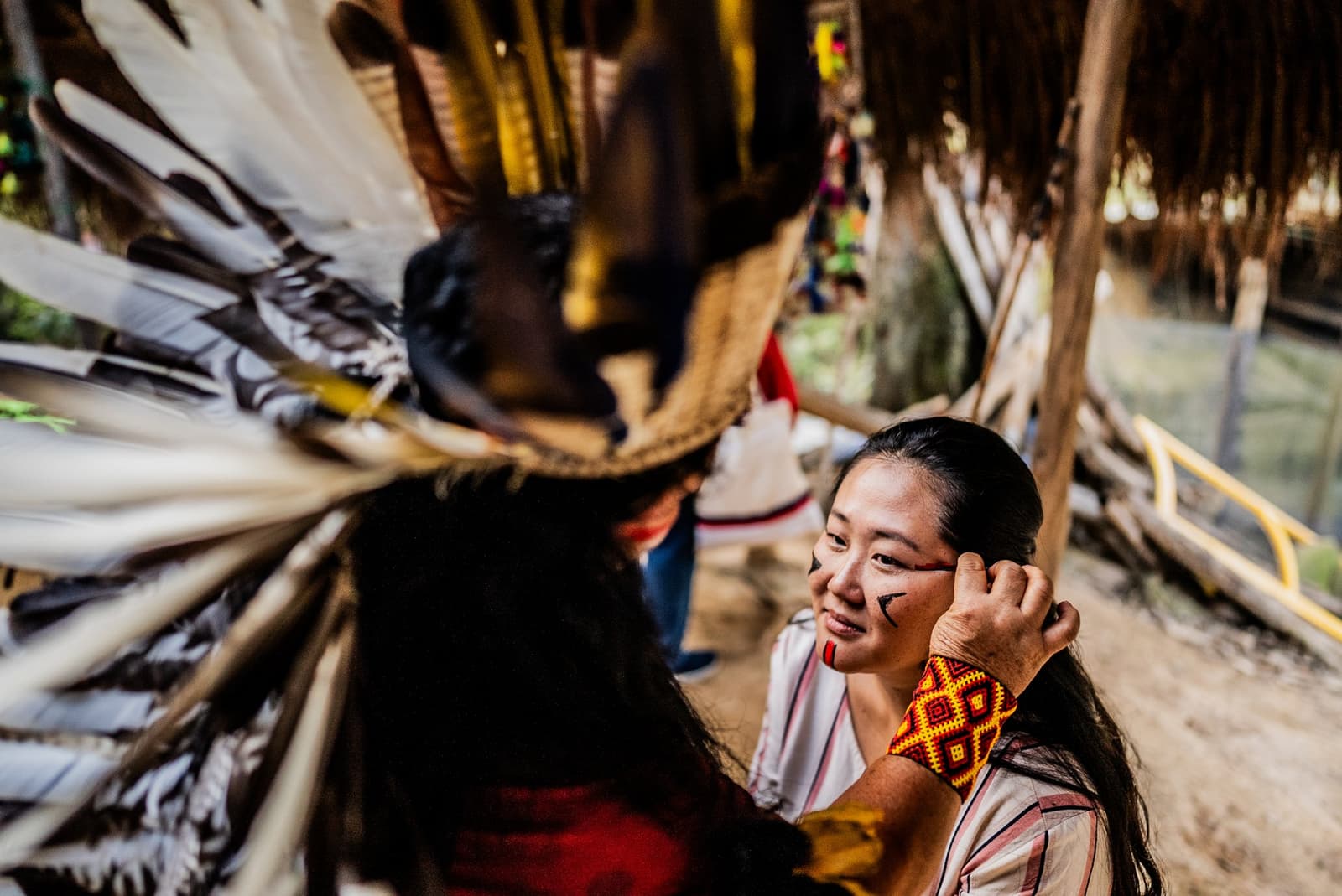 Gesichtsbemalung, Federschmuck und ein Lächeln, das zeigt: dieser Moment ist echt. Photo: FG Trade Eine lächelnde Besucherin mit traditioneller geometrischer Gesichtsbemalung und einem Perlenarmband sitzt einer indigenen Gemeinschaftsperson mit großem Federschmuck gegenüber, während eines Kulturbesuchs auf einer Amazonas-Flusskreuzfahrt in Brasilien