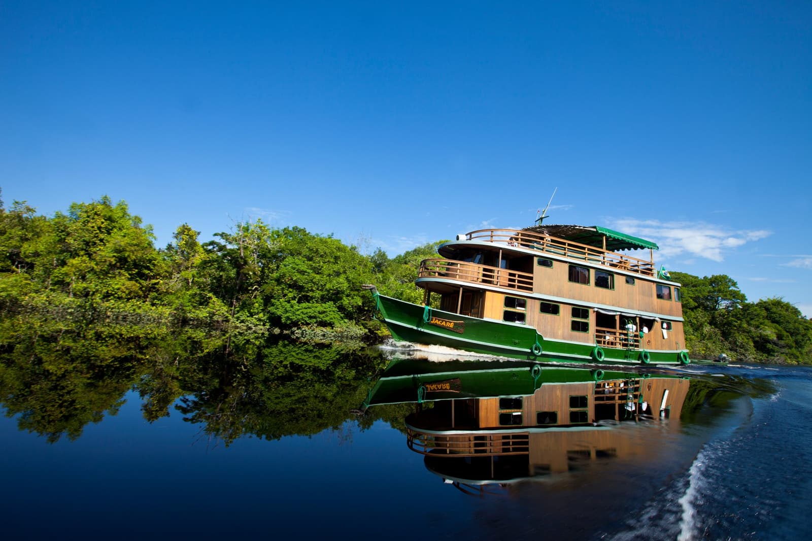 Fünf Nächte an Bord, und der Dschungel gehört ihr. Das Flussboot Jacaré, ein dreistöckiges Holzschiff mit grünem Rumpf, fährt auf einem dunklen, ruhigen Fluss im brasilianischen Amazonas-Gebiet, beidseitig gesäumt von dichtem Regenwald unter strahlend blauem Himmel