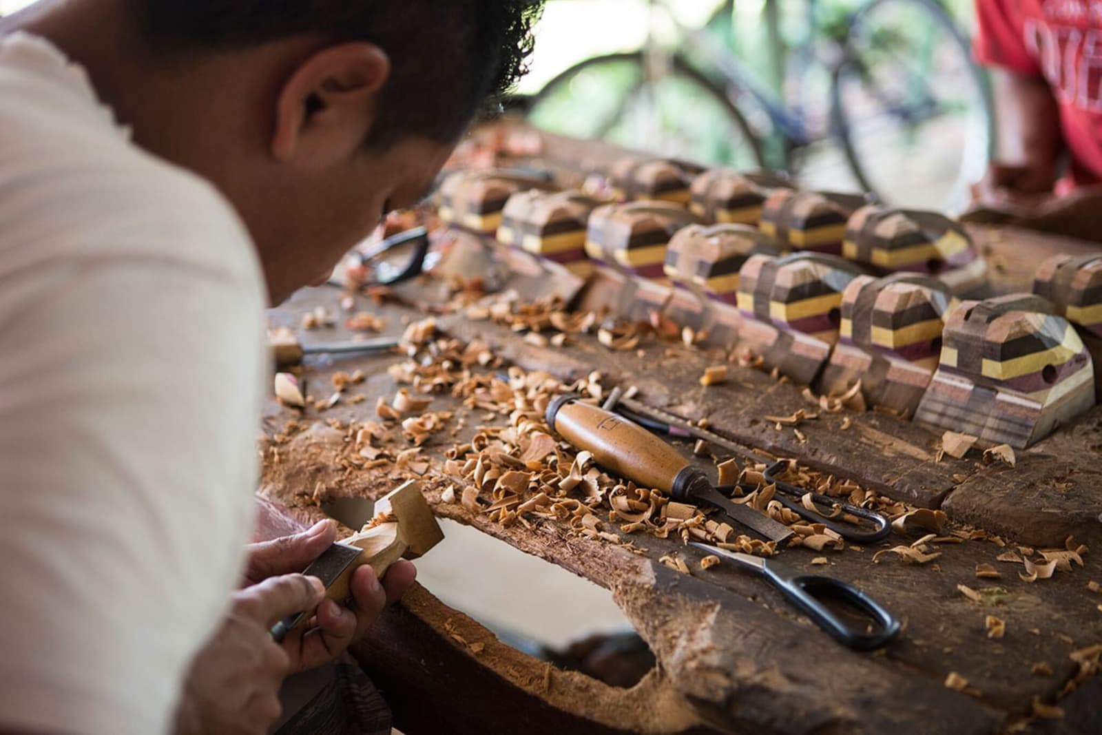 Jahre Übung, ein Messer, und Holzstücke, die man am liebsten alle mitnehmen würde. Ein Handwerker schnitzt von Hand ein kleines Holzstück an einer Werkbank voller Holzspäne, während mehrfarbige Holzfiguren aus laminierten Hölzern aufgereiht daneben liegen