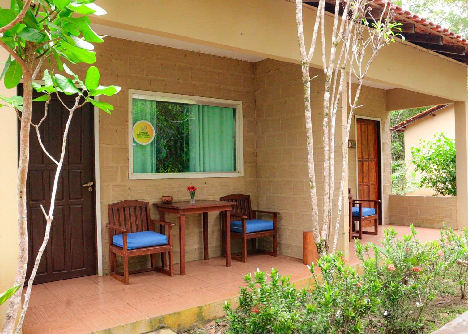 Eine eigene kleine Terrasse, direkt am Rand des Waldes. Überdachte Veranda der Amazon Turtle Lodge mit zwei Holzstühlen mit blauen Kissen und einem kleinen Tisch, umgeben von tropischen Gartenpflanzen und schlanken Bäumen vor einer Steinziegelwand