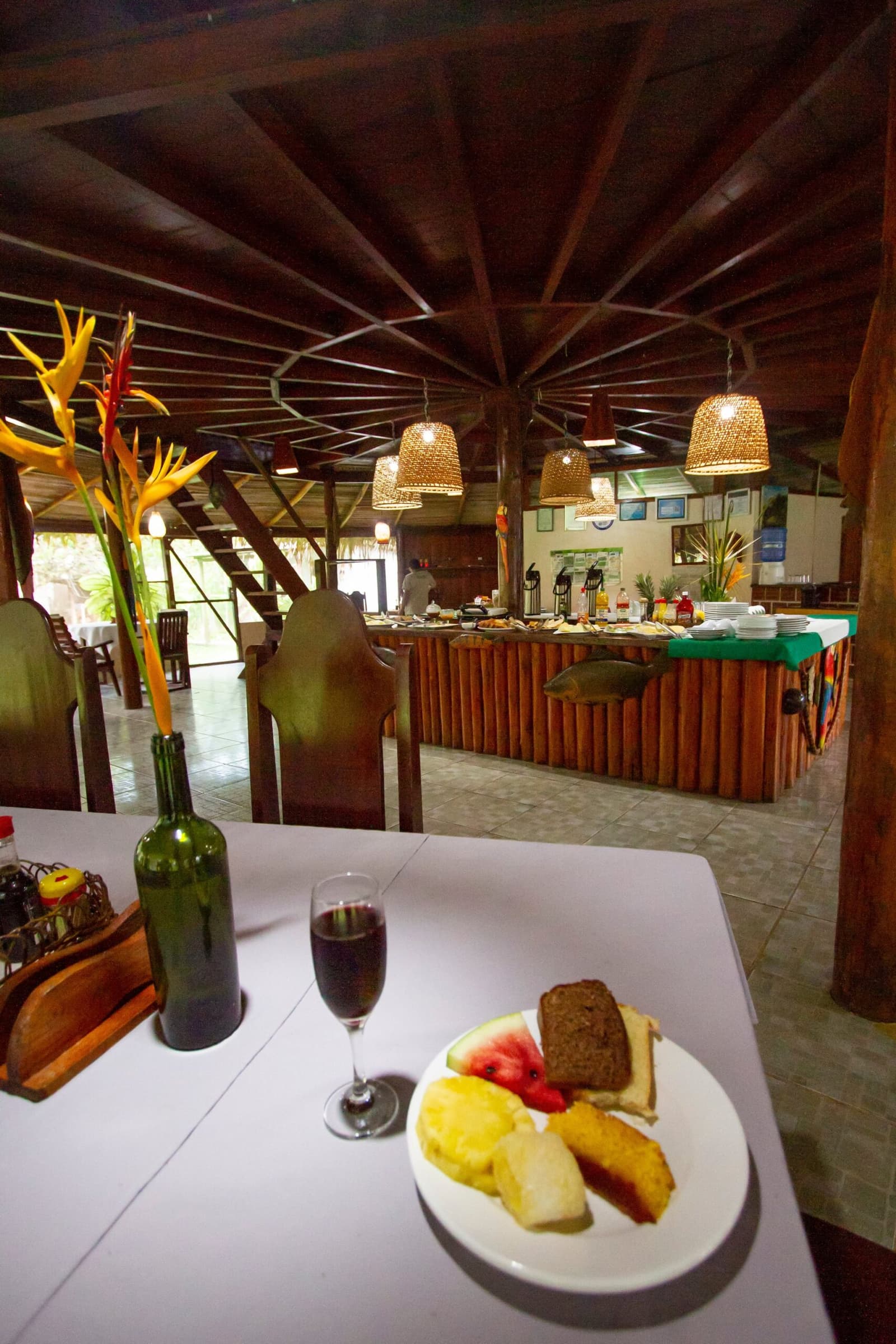 Frühstück bereit, bevor der Regenwald aufwacht. Das Restaurant des Amazon Turtle Lodge zeigt im Vordergrund einen Tisch mit frischen Tropenfrüchten, Brot und Rotwein sowie ein gut bestücktes Buffet, geflochtene Hängelampen und eine markante Holzdecke im Hintergrund