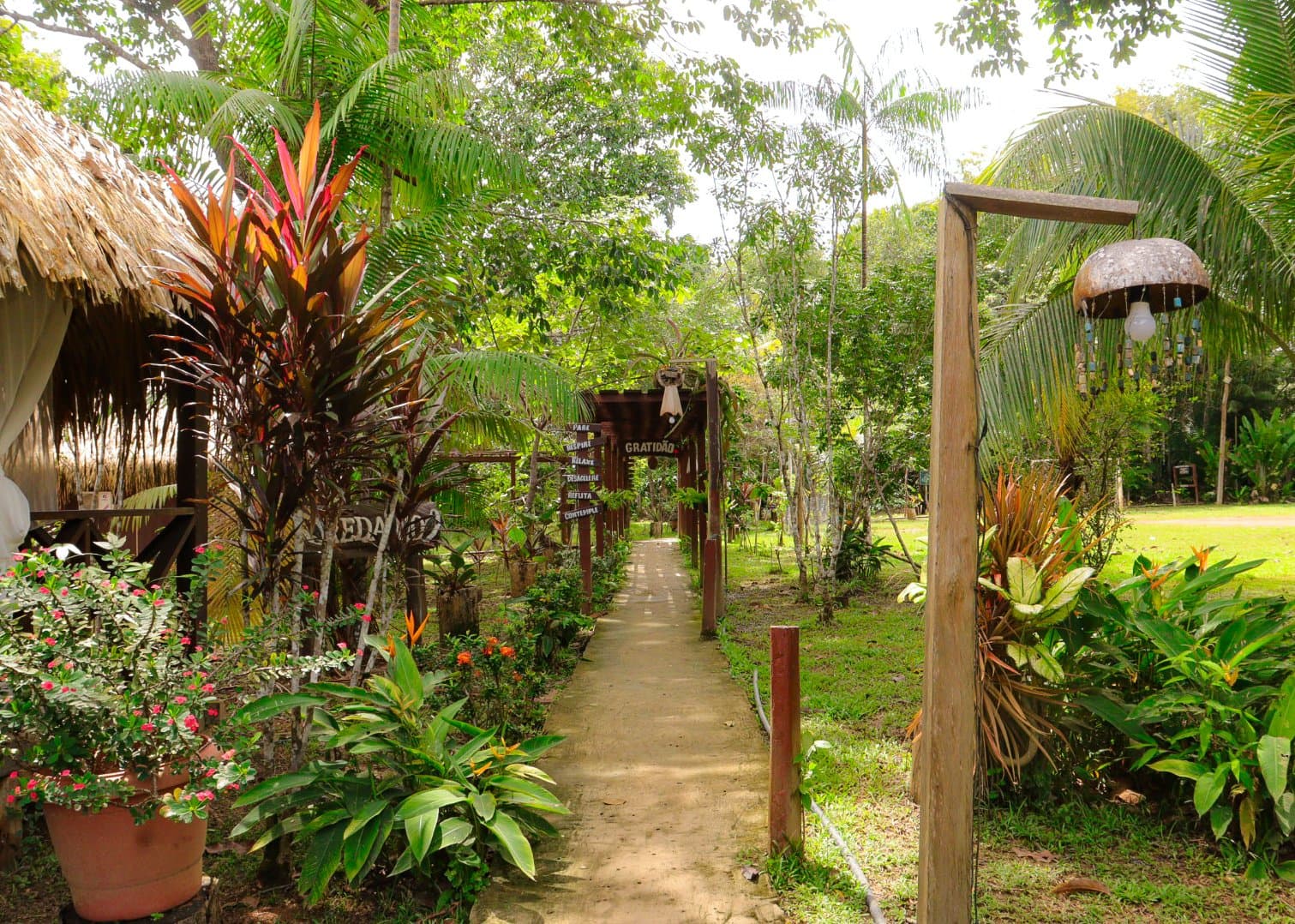 Hier verlangsamt man den Schritt ganz von selbst. Ein schattiger Gartenweg im Amazon Turtle Lodge, gesäumt von bunten Tropenpflanzen, Strohdach-Bungalows und Holzbögen mit portugiesischen Achtsamkeitsschildern