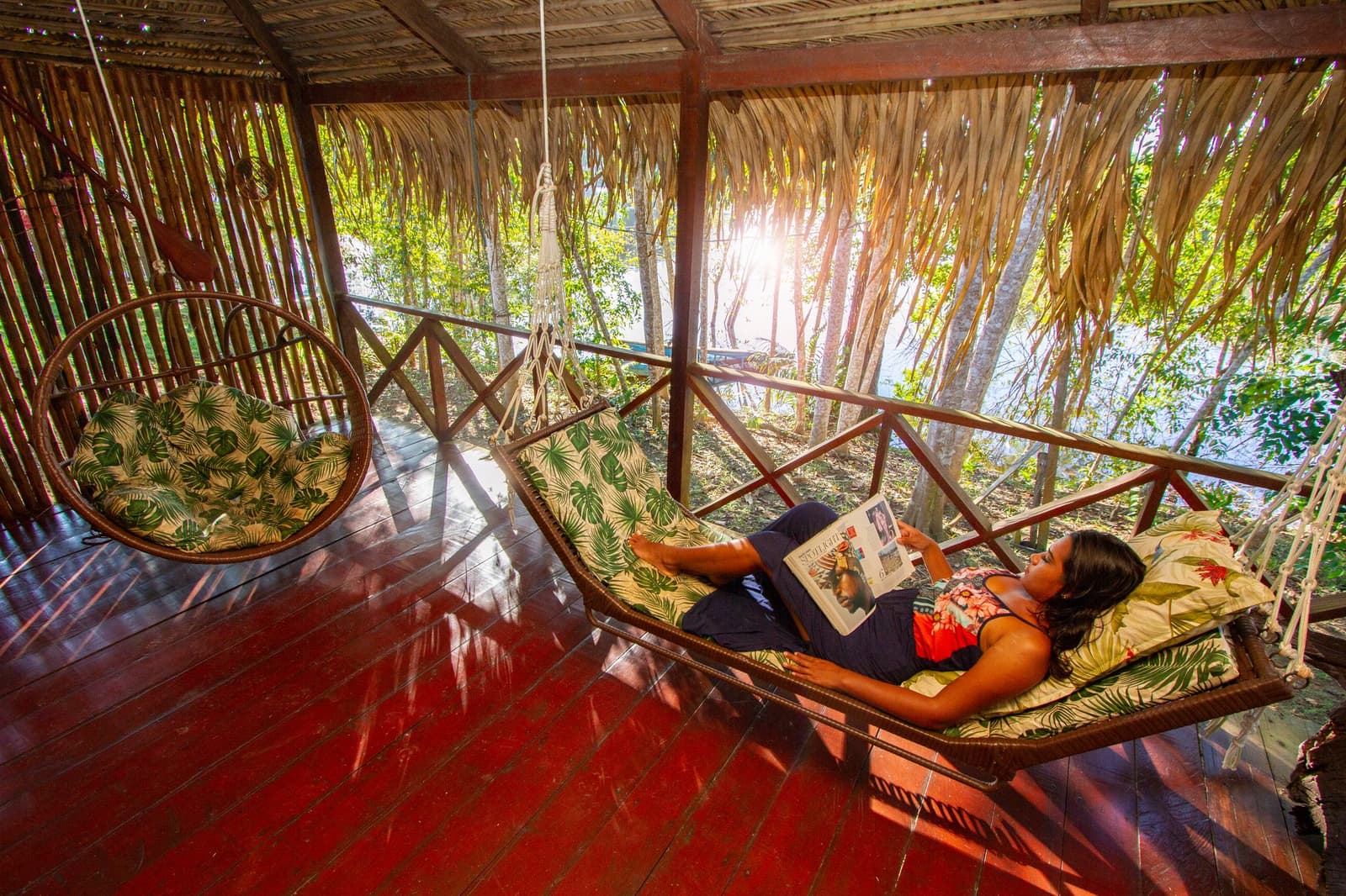 Manchmal braucht der perfekte Amazonas-Nachmittag nicht mehr als eine Hängematte und ein gutes Heft. Eine Gästin liegt lesend in einer gepolsterten Hängematte auf der strohgedeckten Terrasse des Amazon Turtle Lodge, während Sonnenstrahlen durch die tropischen Bäume dahinter fallen