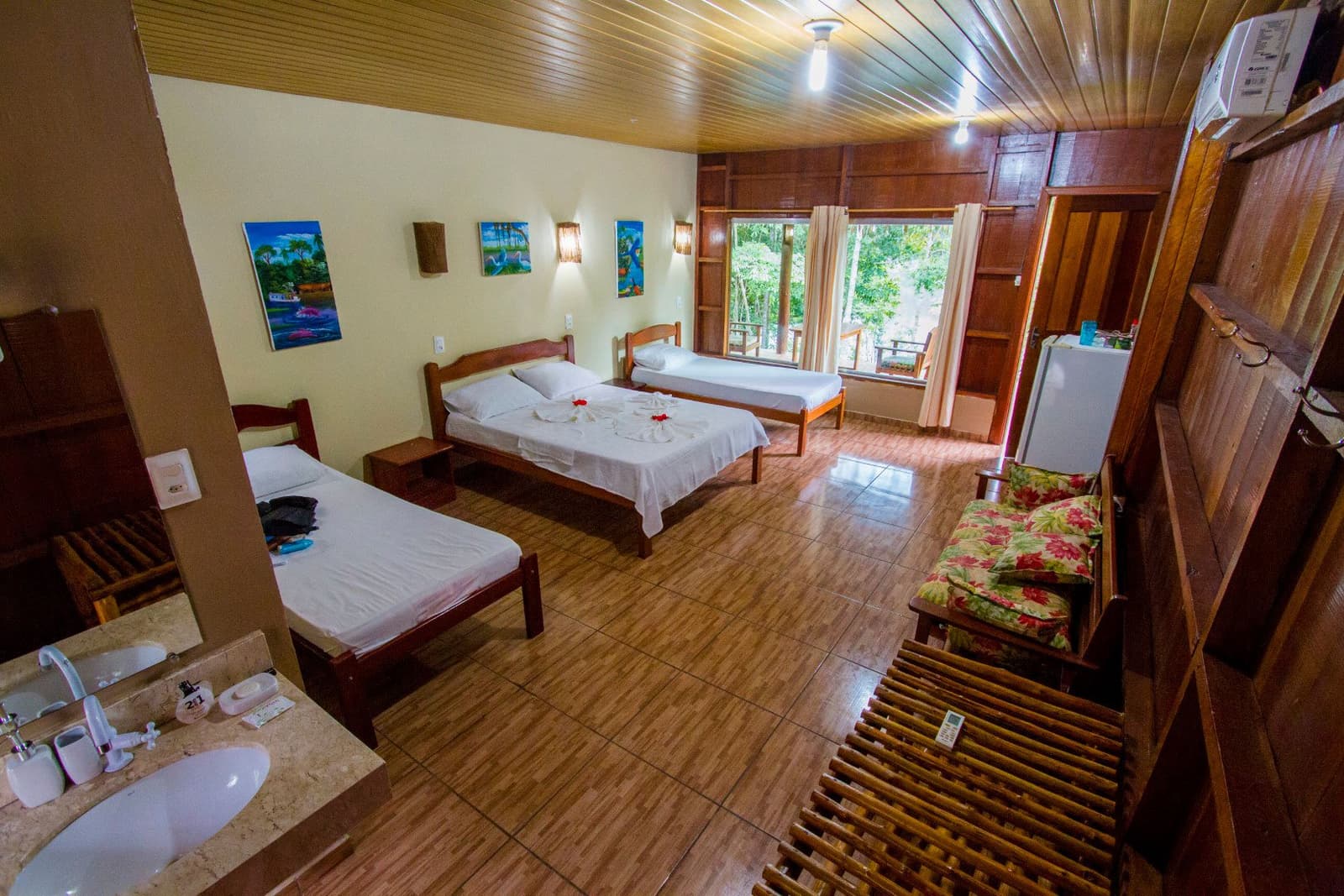 Ordentlich, ruhig und der Dschungel wartet gleich vor der Tür. Geräumiges Dreibettzimmer im Amazon Turtle Lodge mit drei Holzbetten in weißer Bettwäsche, tropischen Gemälden an cremefarbenen Wänden und einem Fenster mit Blick in den Regenwald