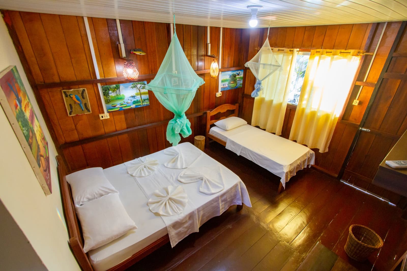 Genug Komfort, um den Dschungel draußen zu lassen. Holzgetäfeltes Doppelzimmer im Amazon Turtle Lodge mit einem Doppelbett und einem Einzelbett, grünen und weißen Moskitonetzen, gefalteten Handtüchern auf weißer Bettwäsche und bunten Regenwald-Gemälden an den Wänden
