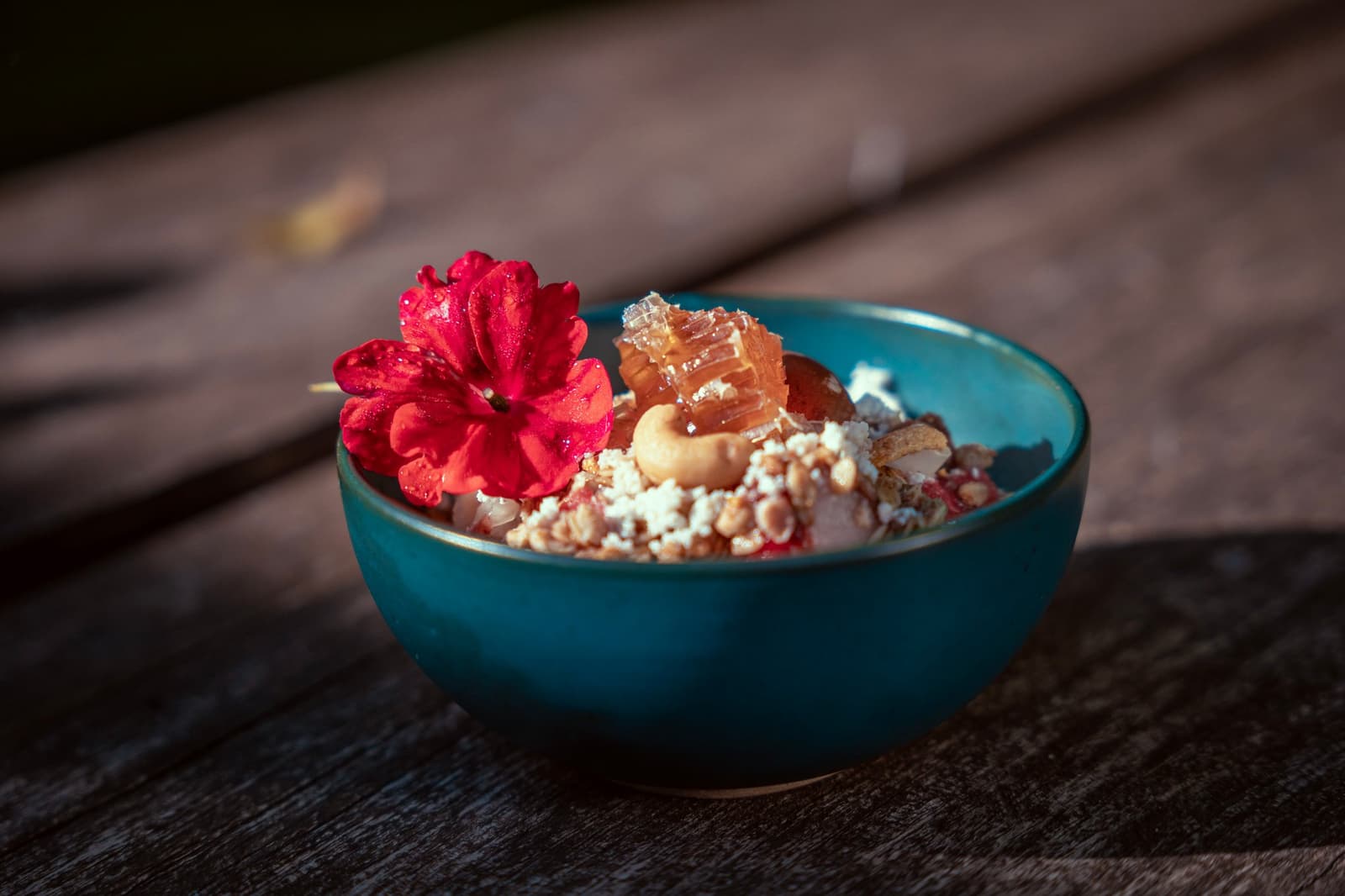 Le petit-déjeuner mérite sa fleur. À table avant que les oiseaux ne s'envolent. Un bol en céramique bleu canard rempli de granola, de noix de cajou, de rayon de miel et de fromage émietté, garni d'une fleur rouge, posé sur une table en bois rustique dans un lodge amazonien