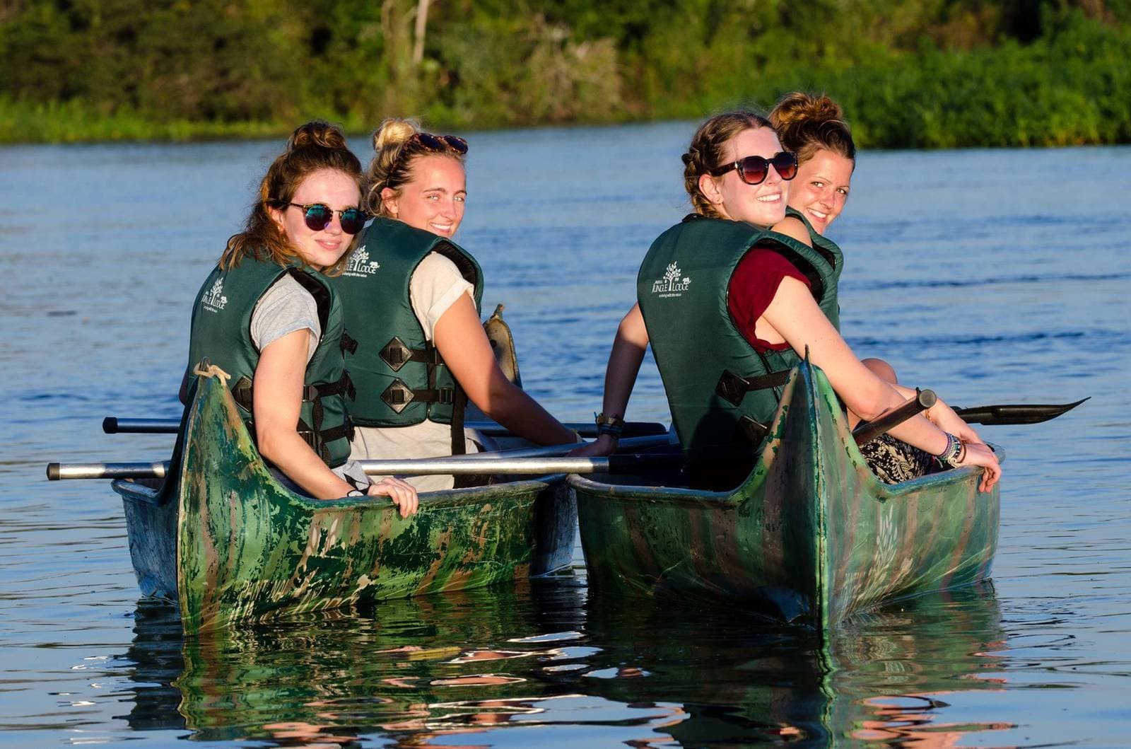 Good company, calm water, and nowhere better to be. Four smiling women in green life jackets paddling a canoe on a calm river during a budget Pantanal tour, dense green forest lining the far bank