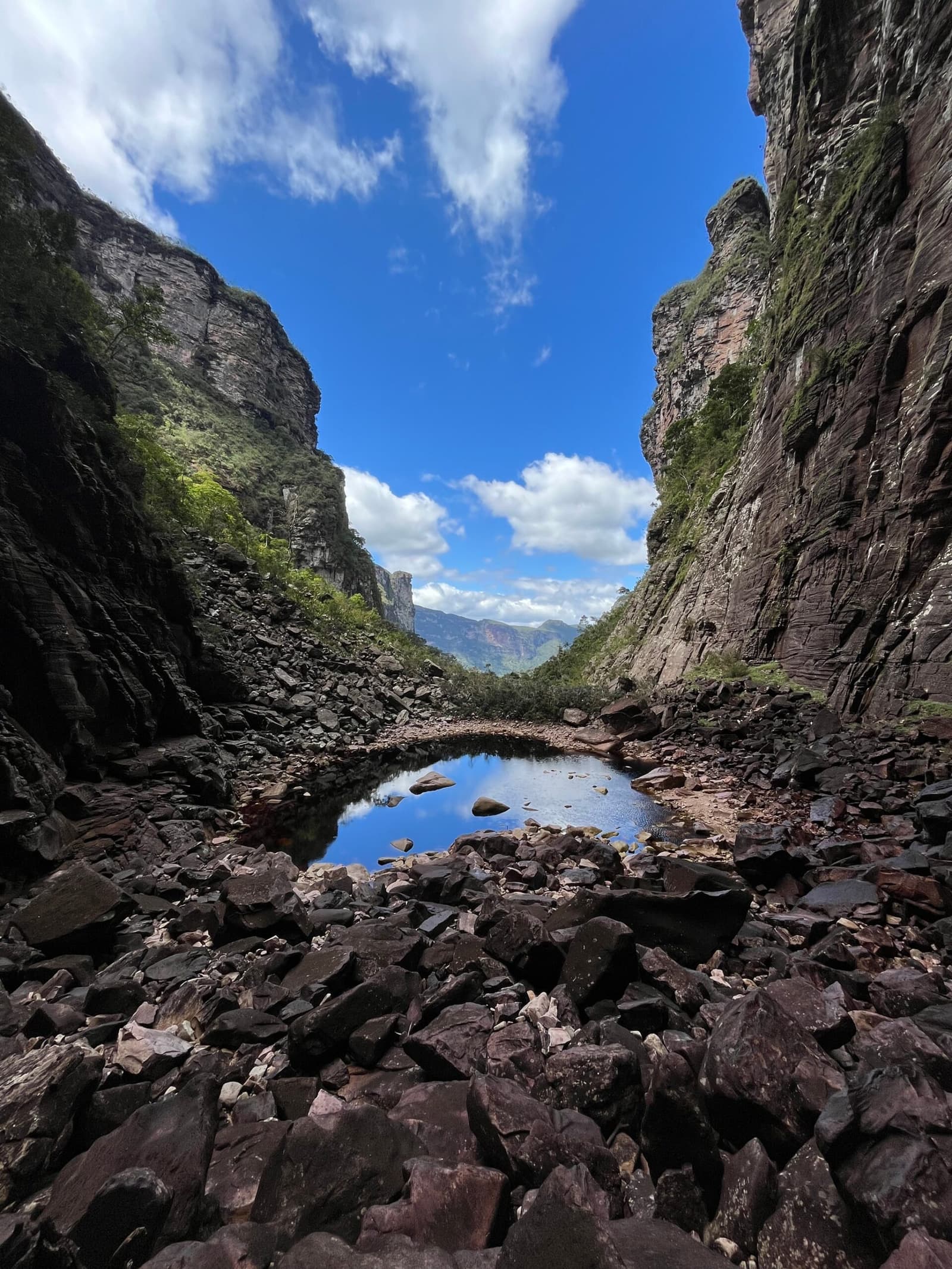 Ein Stück Himmel, eingefasst in Stein und für einen Moment angehalten. Photo: Lucas Neves Eine enge Schlucht auf dem Chapada-Diamantina-Trek mit hohen Sandsteinwänden, die einen stillen Felsenpool flankieren, der den blauen Himmel spiegelt, umgeben von dunklen Felsblöcken mit fernen grünen Bergen im Hintergrund
