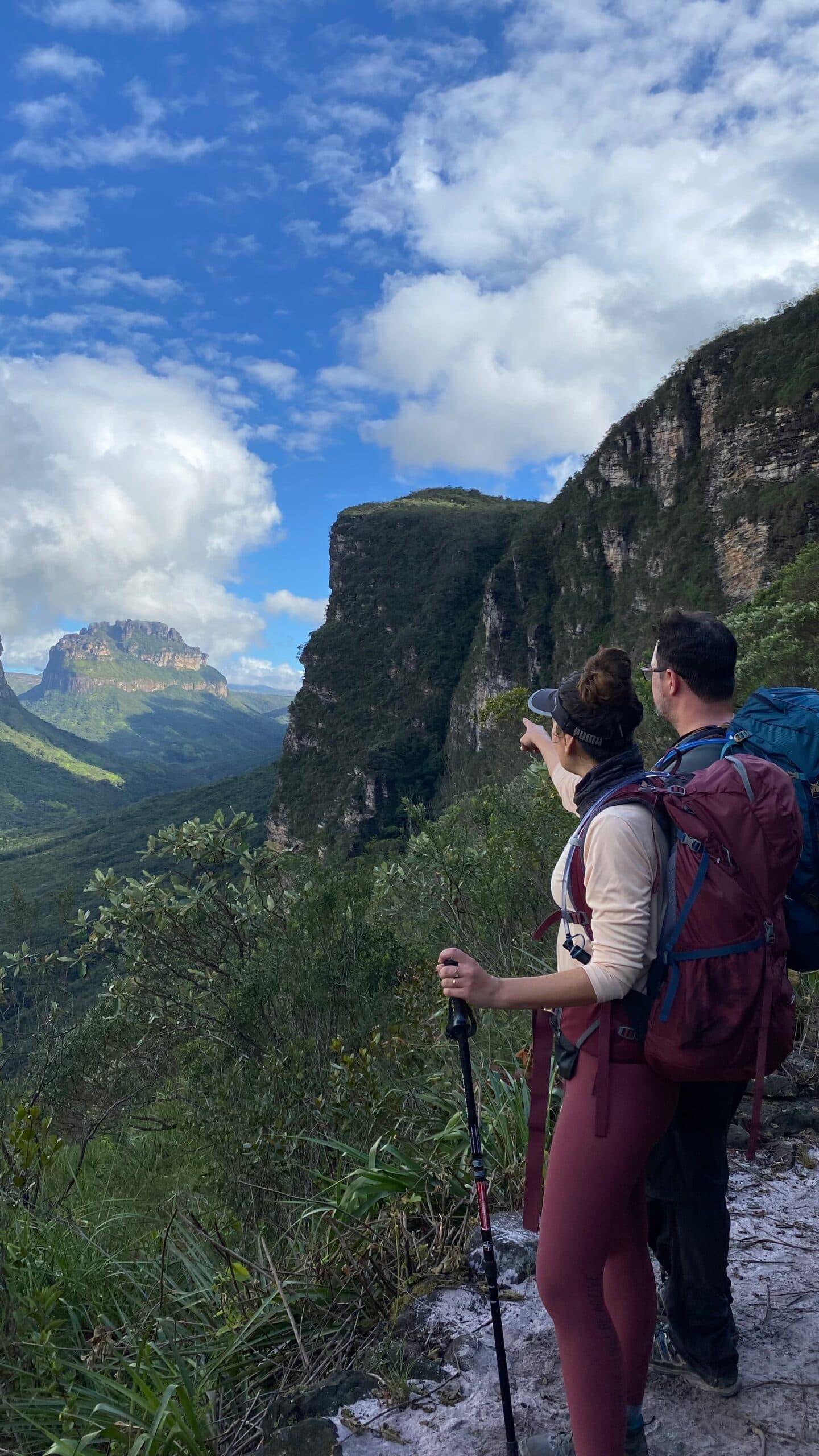 Eine Hand zeigt, und das Tal ergibt plötzlich seinen Sinn. Photo: Lucas Neves Zwei Wanderer mit Rucksäcken und Trekkingstöcken machen auf einem Felsvorsprung beim Chapada-Diamantina-Trek Halt; eine Person zeigt auf das weite Canyontal mit tafelförmigen Sandsteinmesas unter einem teilweise bewölkten Himmel