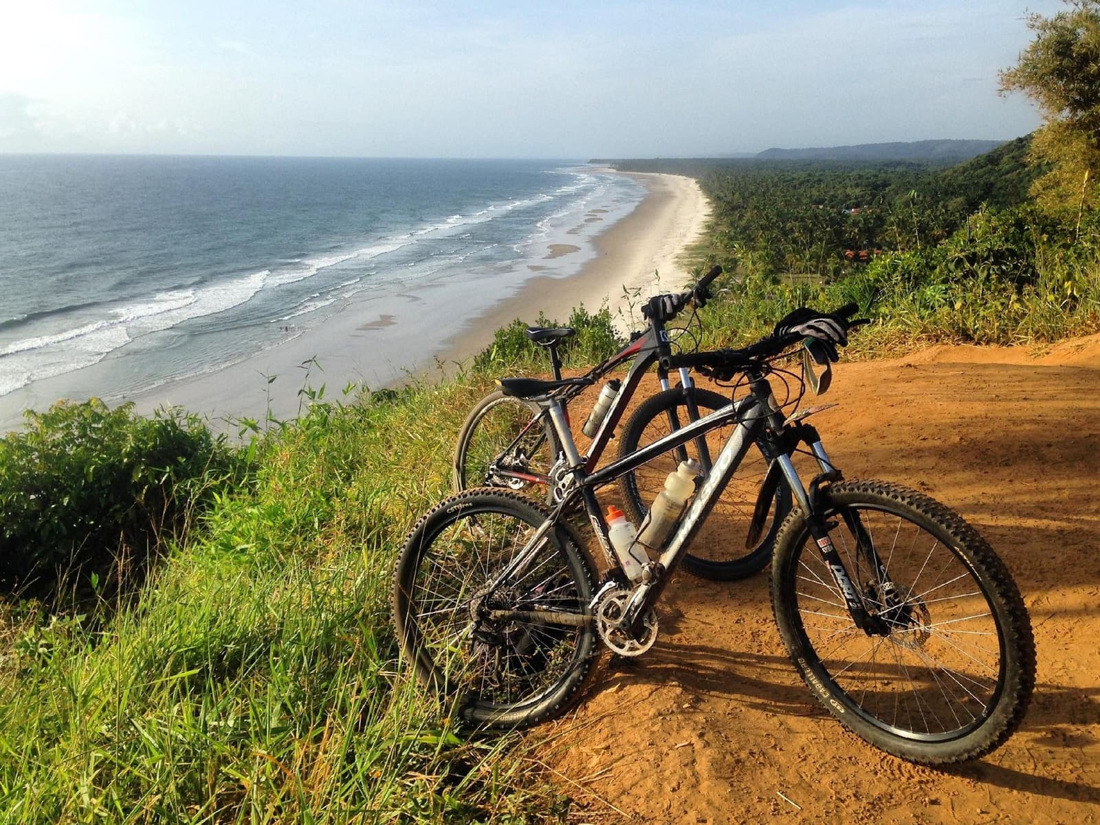 Zwei Räder, eine Aussicht, und ein Strand ohne Ende. Zwei Mountainbikes auf einem roten Feldweg am Klippenrand bei einer Bahia-Radtour, darunter ein langer leerer Strand, Atlantikwellen und dichter Küstenwald
