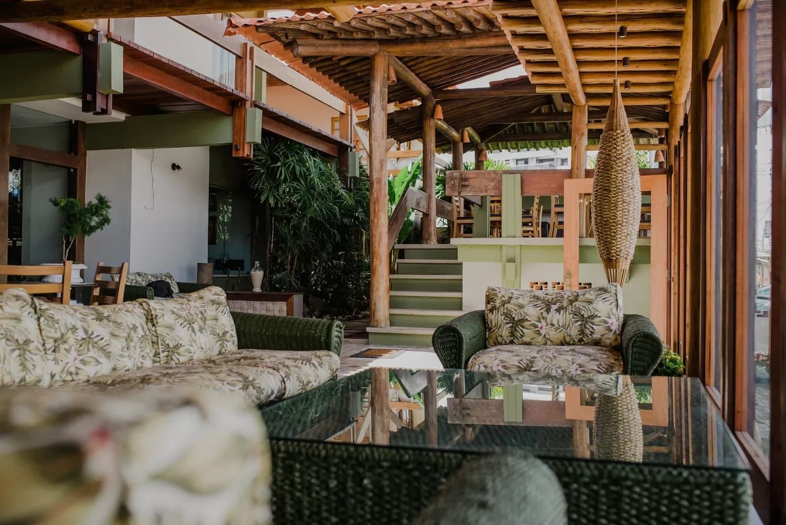 Sofas so bequem, dass man den Wecker fast vergisst. Offene Lounge mit Korbsofas und geblümten Kissen unter einem rustikalen Holzbalken-Dach, Zimmerpflanzen und eine Treppe im Hintergrund in einer Pousada auf der Radroute Bahia Kakaoroute